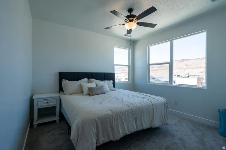 Bright secondary bedroom featuring large dual windows with expansive natural light, a ceiling fan with integrated lighting, dark-finish flooring, and a white nightstand