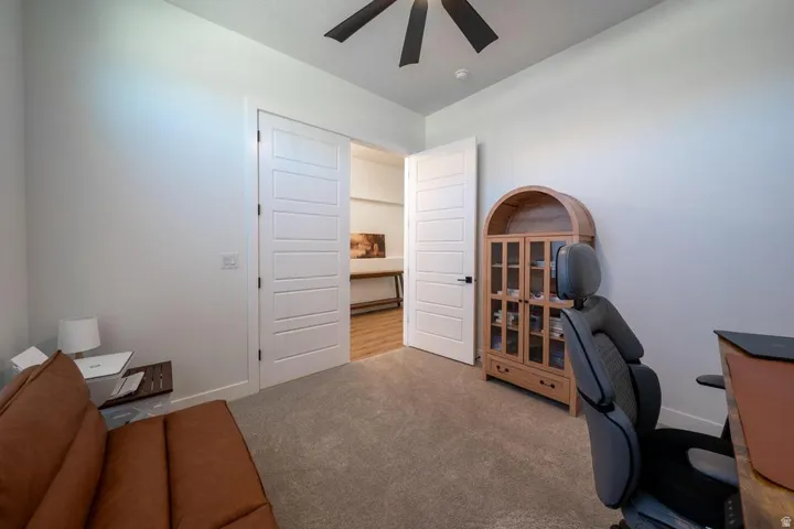 Versatile multi-purpose room featuring neutral carpet flooring, ceiling fan with integrated lighting, dual white panel doors with transom windows, and built-in wood-finish cabinetry with glass-front display doors