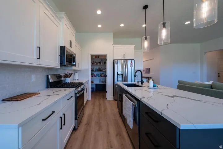 Modern galley kitchen featuring white shaker cabinetry, quartz countertops with waterfall edge detailing, stainless steel appliances, subway tile backsplash, wood-finish flooring, pendant lighting, and a contrasting dark blue island with integrated sink a