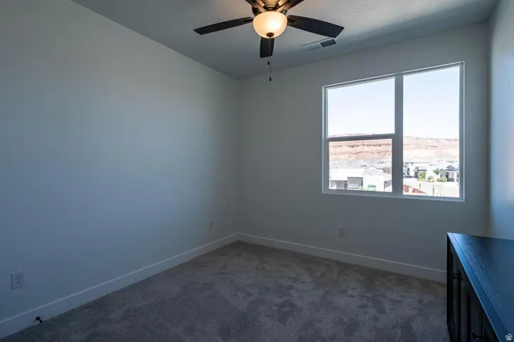 Spacious room featuring a large window, neutral wall tones, gray carpet, white baseboards, and a ceiling fan with integrated lighting