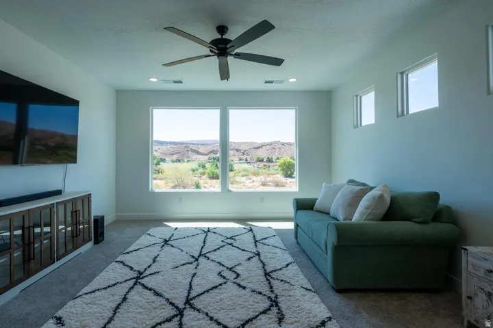 Bright upper-level room featuring expansive windows framing scenic hillside vistas, ceiling fan with dark finish, geometric patterned area rug in neutral tones, green upholstered seating, built-in cabinetry with wood-finish doors, recessed lighting, and n