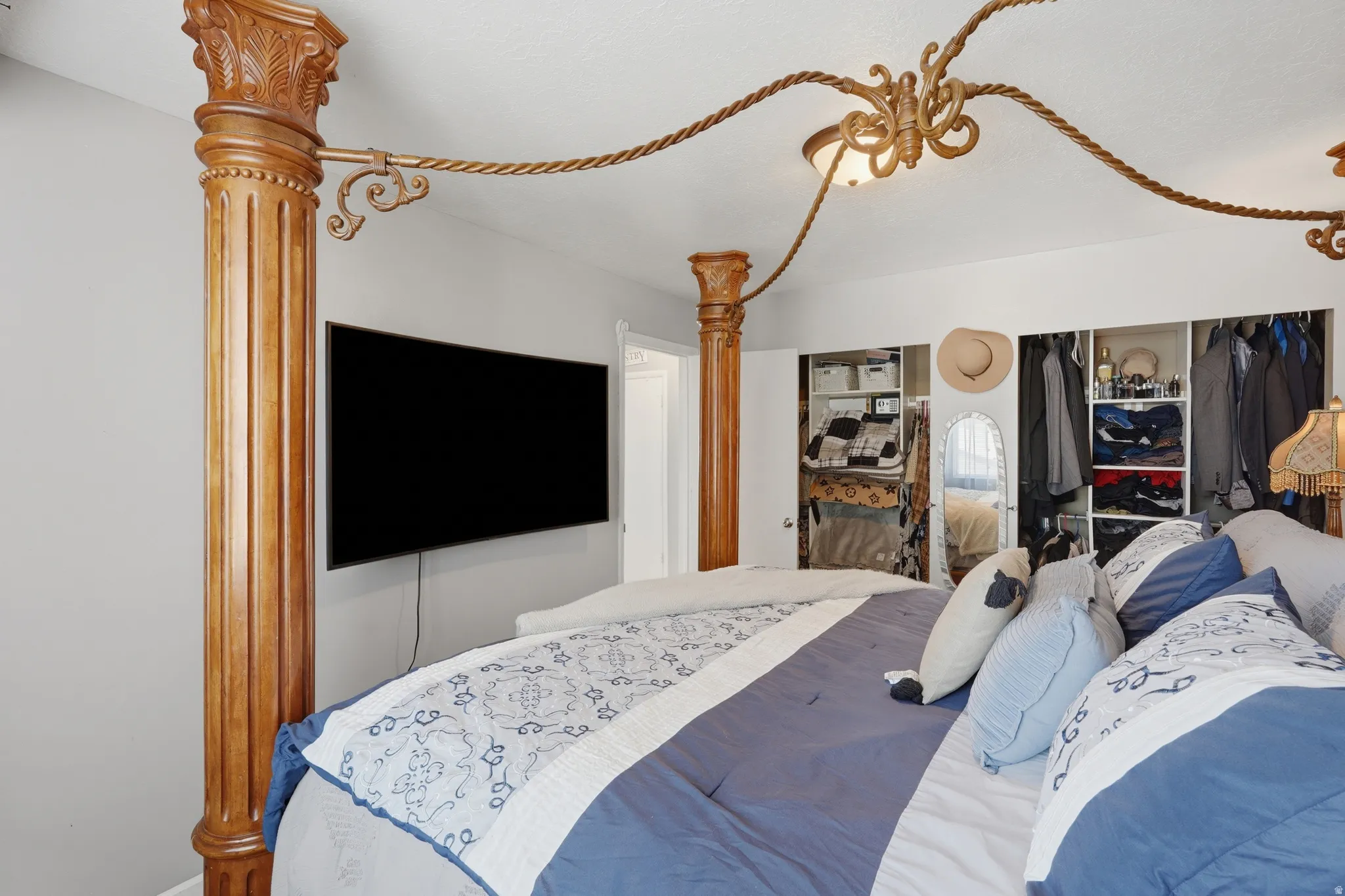 Elegant primary bedroom features a striking four-poster bed with ornamental carved wood details and rope swag accents, wall-mounted flat-screen television, mirrored closet doors with built-in storage, and crisp blue and white bedding in a bright, neutral