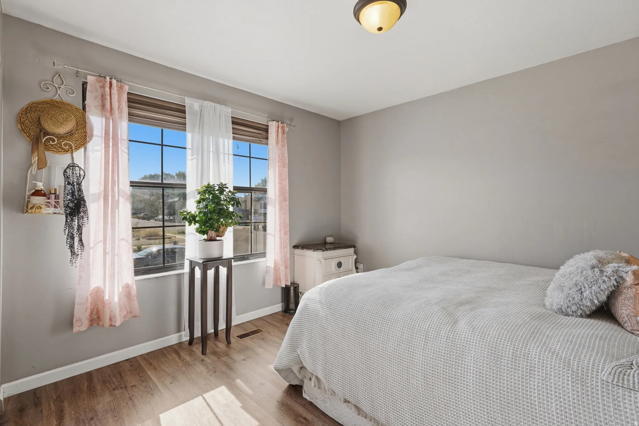 Bright bedroom featuring expansive windows with pink curtains framing scenic landscape views, hardwood flooring, neutral gray walls, ceiling-mounted lighting fixture, and ample natural light throughout the space