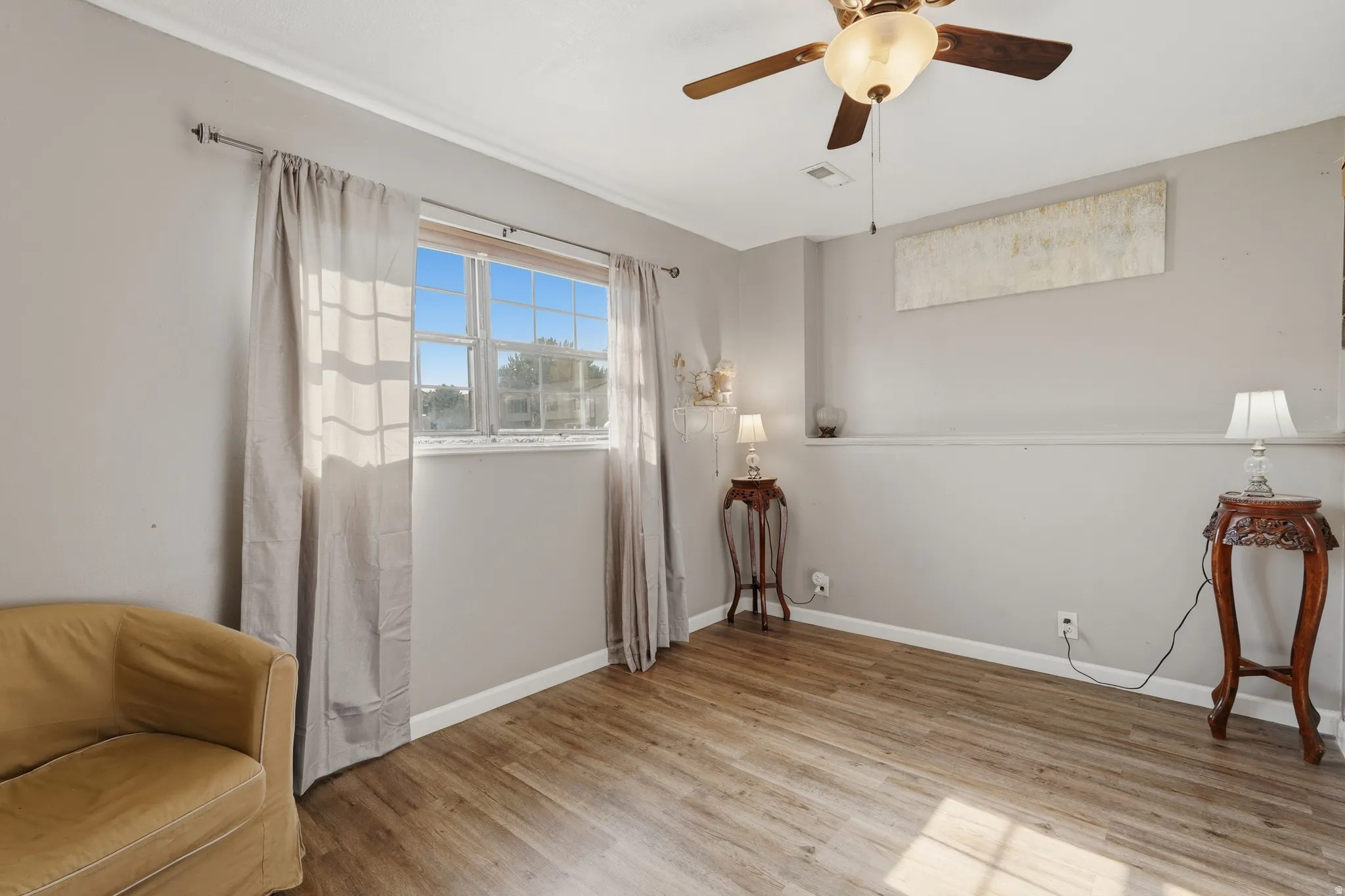 Bright, light-filled bedroom featuring wood-finish flooring, a ceiling fan with integrated lighting, and large window with sheer curtains allowing natural light