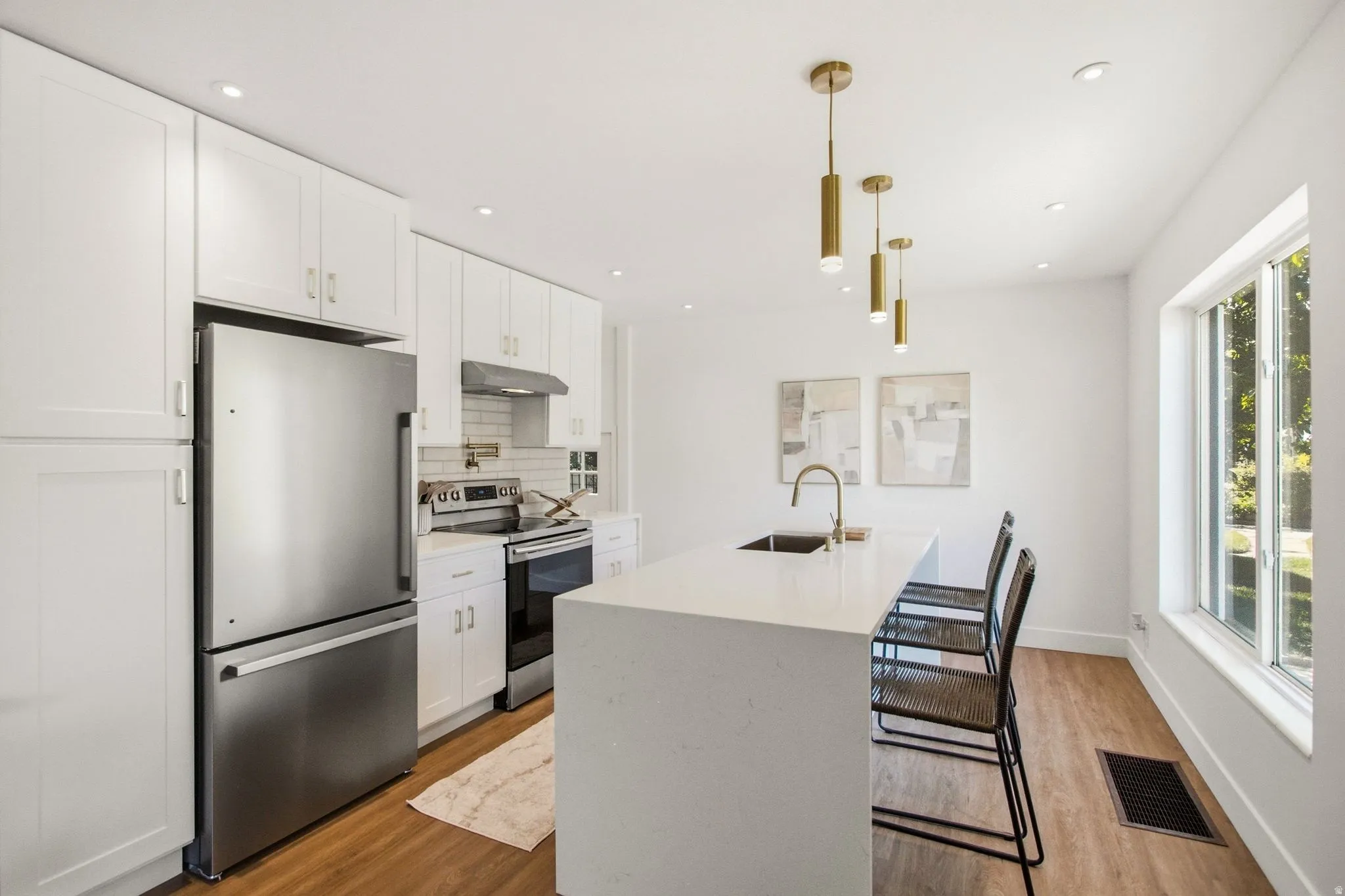 Modern kitchen featuring white shaker cabinetry, stainless steel appliances including a French-door refrigerator and gas range, a waterfall-edge white quartz island with seating for two, warm wood-finish flooring, recessed ceiling lighting, and brass pend