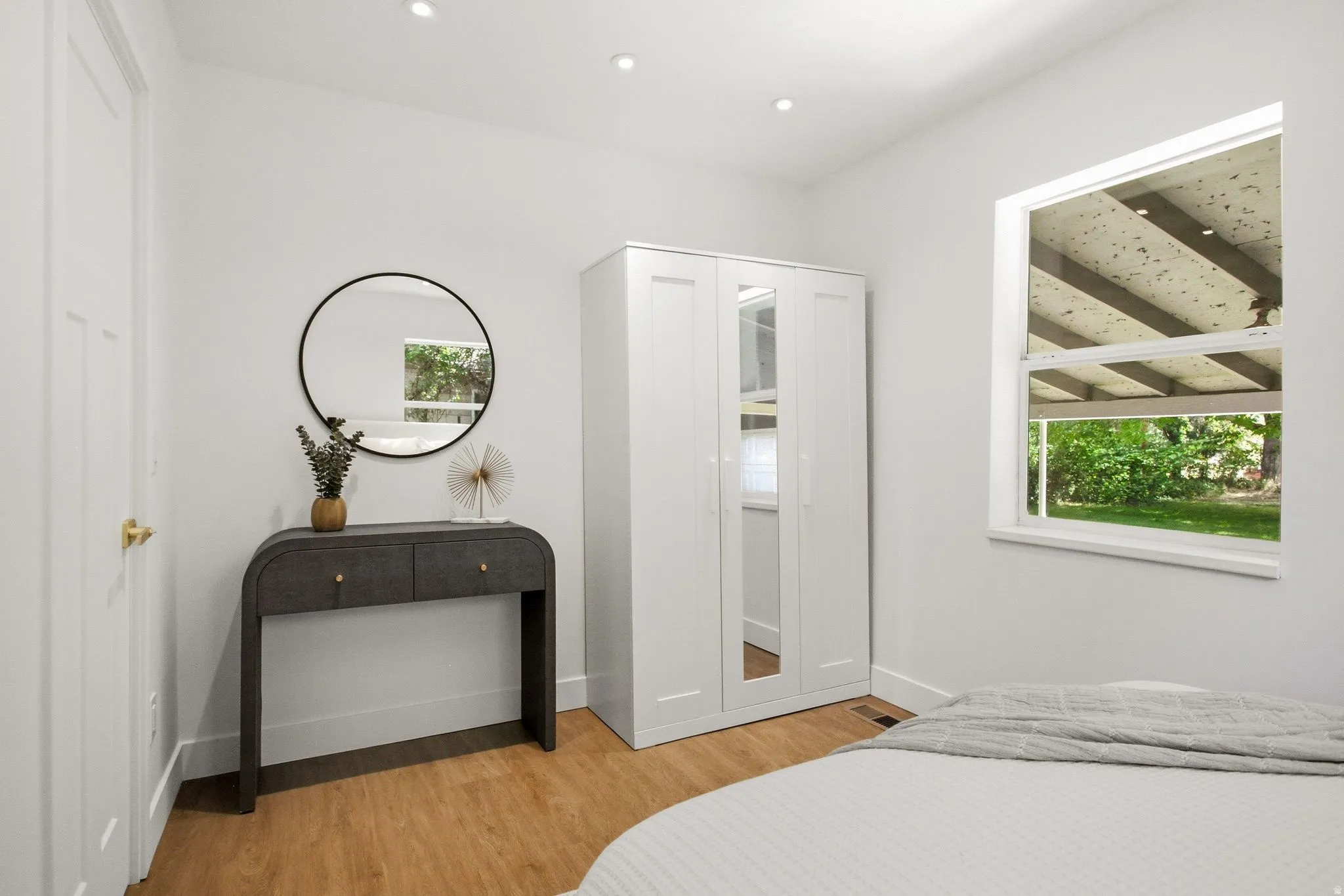 Bright, airy bedroom featuring warm wood-finish flooring, recessed ceiling lighting, and a large operable window with exterior wooden pergola visible