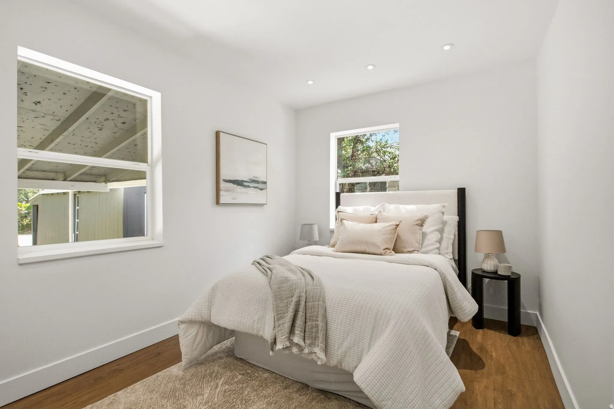 Serene bedroom showcasing warm wood-finish flooring, white painted walls, and abundant natural light from dual windows