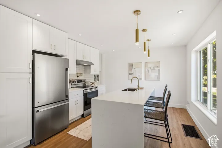 Modern kitchen featuring white shaker cabinetry, stainless steel appliances including a French-door refrigerator and gas range, a waterfall-edge white quartz island with seating for two, warm wood-finish flooring, recessed ceiling lighting, and brass pend