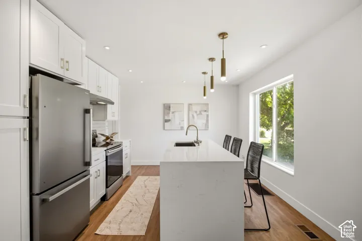 Modern kitchen featuring white cabinetry with brushed brass hardware, stainless steel appliances including a French-door refrigerator and slide-in range, white quartz waterfall island countertop, pendant lighting with brass accents, wood-finish flooring,