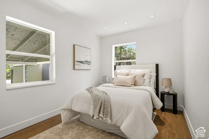 Serene bedroom showcasing warm wood-finish flooring, white painted walls, and abundant natural light from dual windows