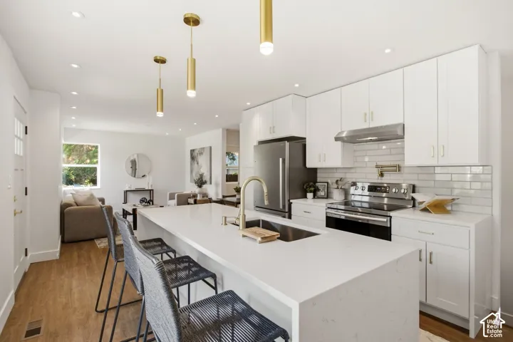 Contemporary kitchen featuring white cabinetry, a large waterfall-edge quartz island with seating for multiple, stainless steel appliances including a gas range and under-counter refrigeration, subway tile backsplash, and pendant lighting