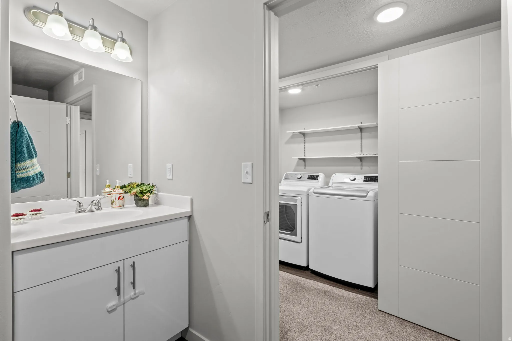 Convenient laundry room with side-by-side washer and dryer, white cabinetry with ample storage, recessed lighting, and direct access to the adjoining bathroom featuring a vanity sink, chrome fixtures, and mirror with integrated lighting