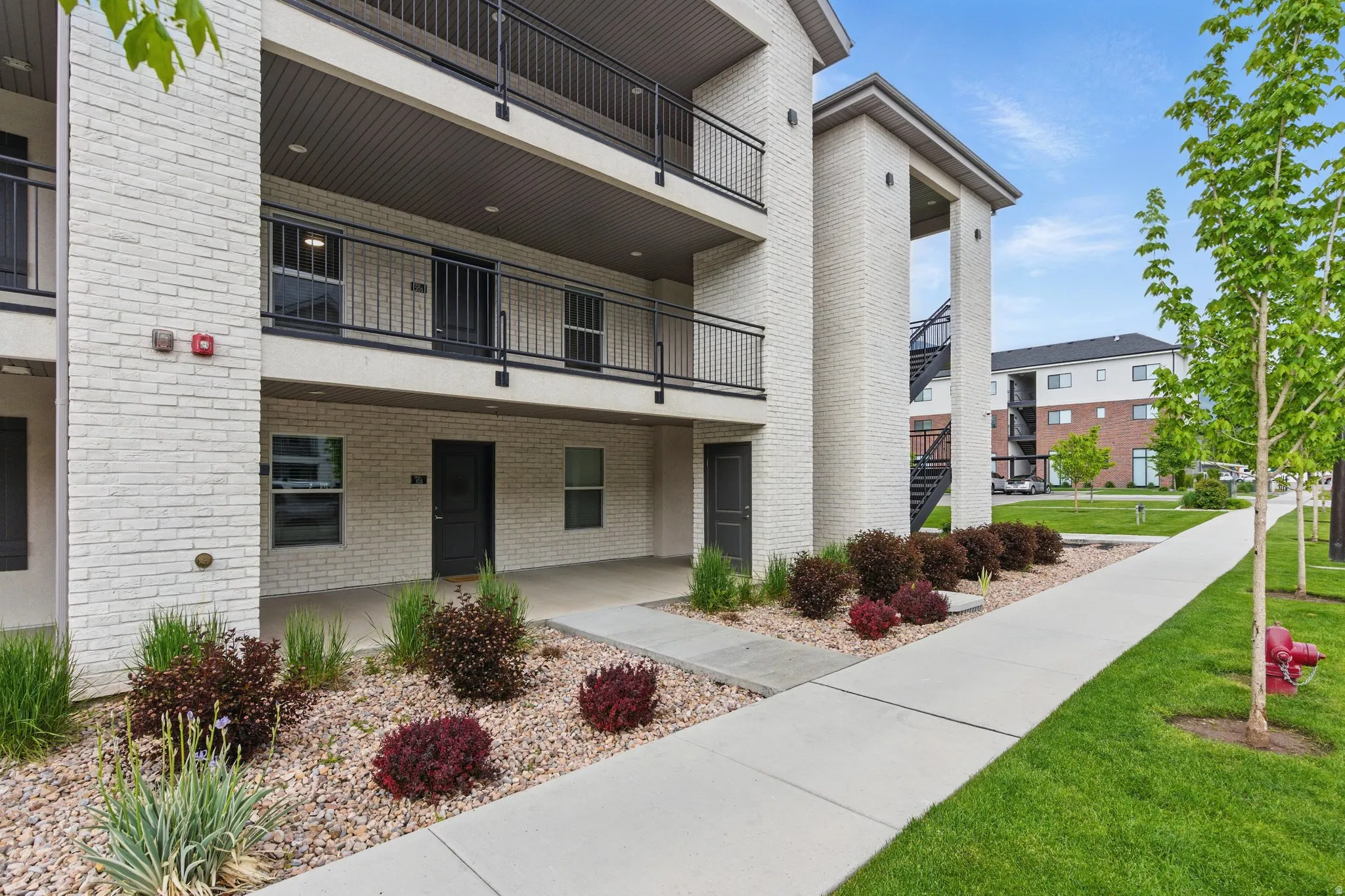 Multi-level building exterior featuring light-colored brick, dark balconies with metal railings, and a covered entry area