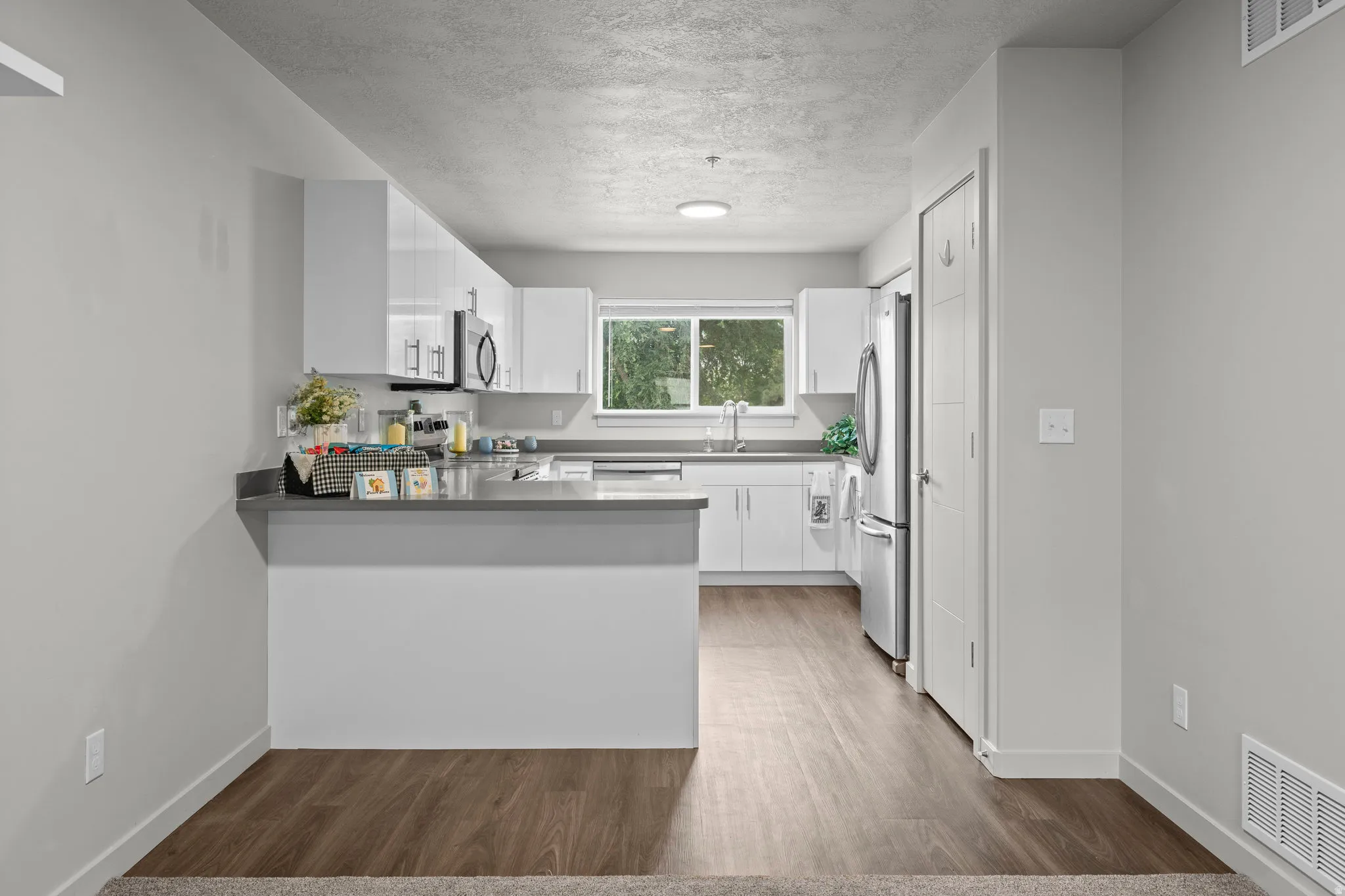 Open-concept kitchen and dining area featuring white cabinetry with a gray quartz countertop island, stainless steel appliances, wood-finish flooring throughout, and abundant natural light from large windows