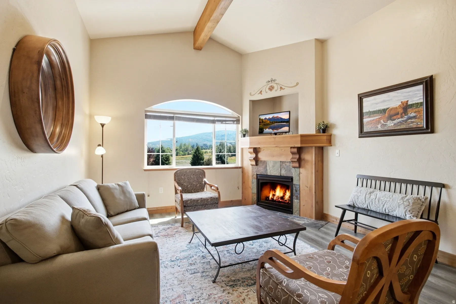 Arched window providing natural light, vaulted ceiling with exposed wood beam, natural stone fireplace with wood mantel, built-in media niche, and wood-finish flooring