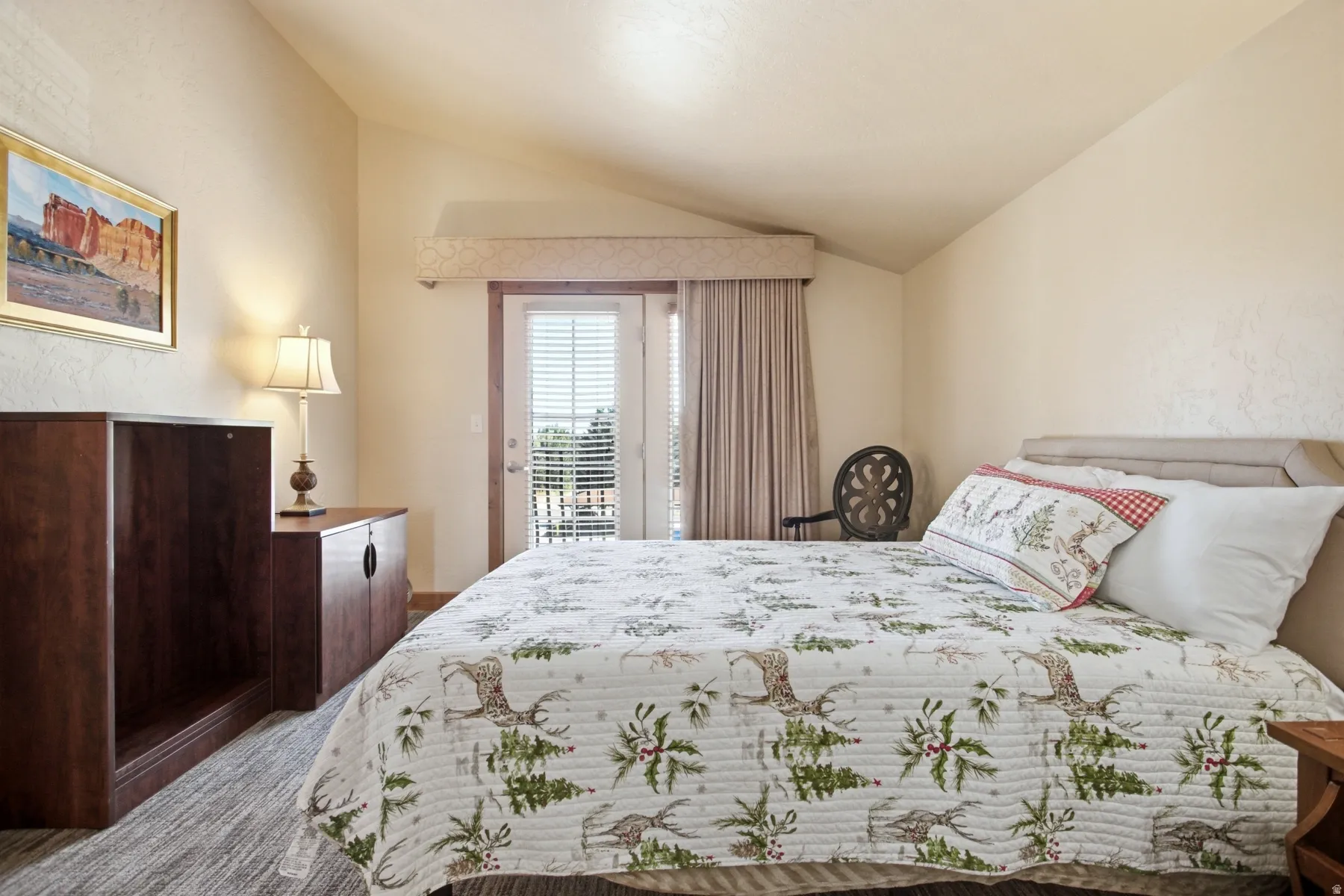 Spacious bedroom featuring a wood-finish dresser, recessed ceiling lighting, glass sliding door access to a private balcony with operable curtains, plush bedding with botanical print, and coordinating decorative pillows