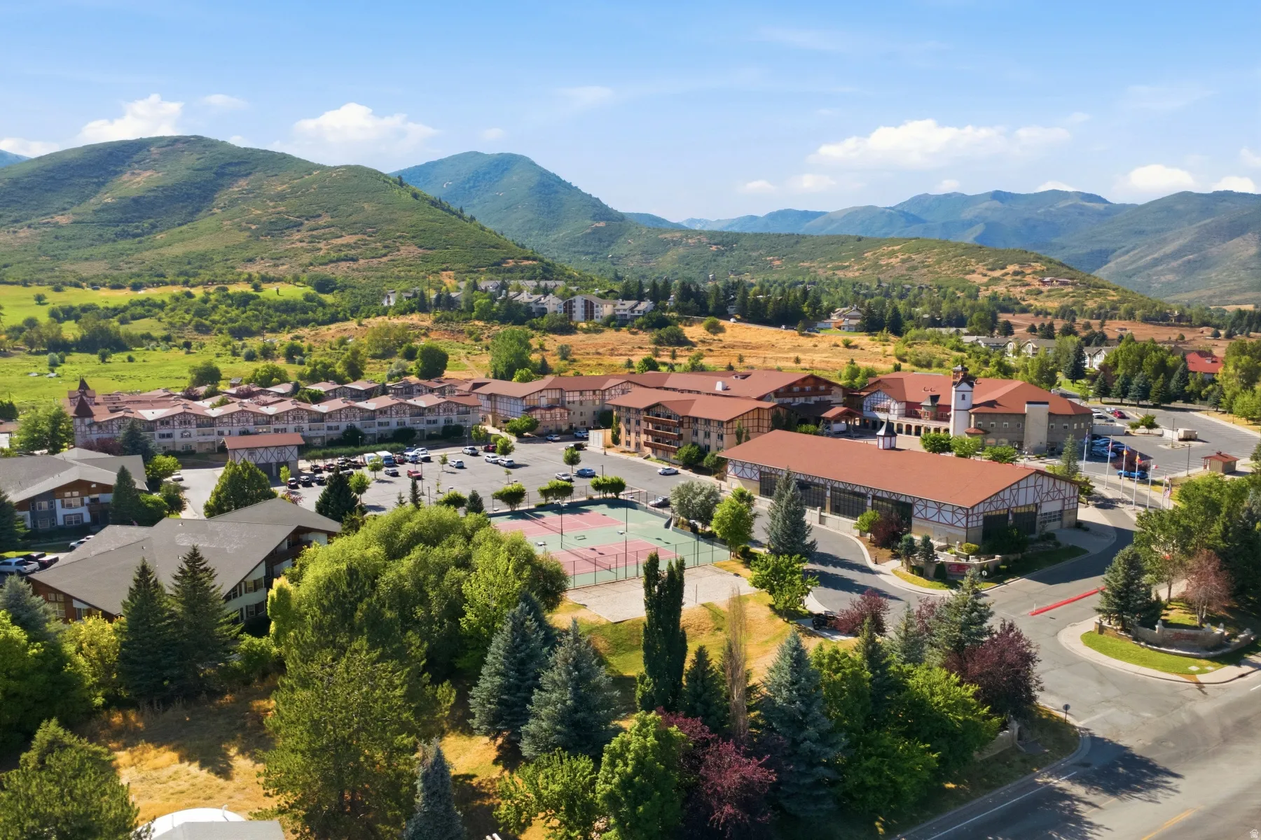 Expansive community featuring multiple buildings with gabled roofs, a central tennis court, and paved parking areas