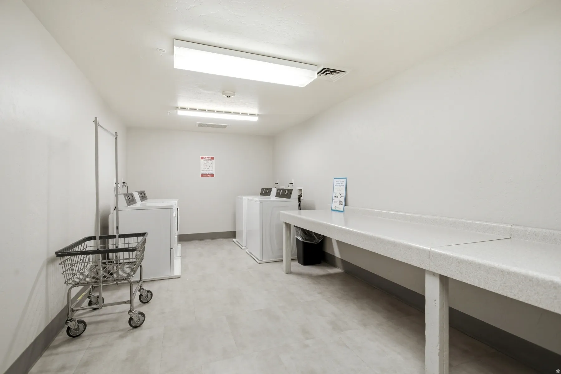 Dedicated laundry room featuring commercial-grade washer and dryer units, generous countertop workspace with stainless steel sink and faucet, bright overhead LED lighting, and neutral tile flooring throughout