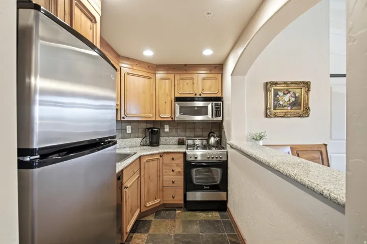 Compact kitchen featuring stainless steel appliances, wood cabinetry, stone tile flooring, and light-toned countertops