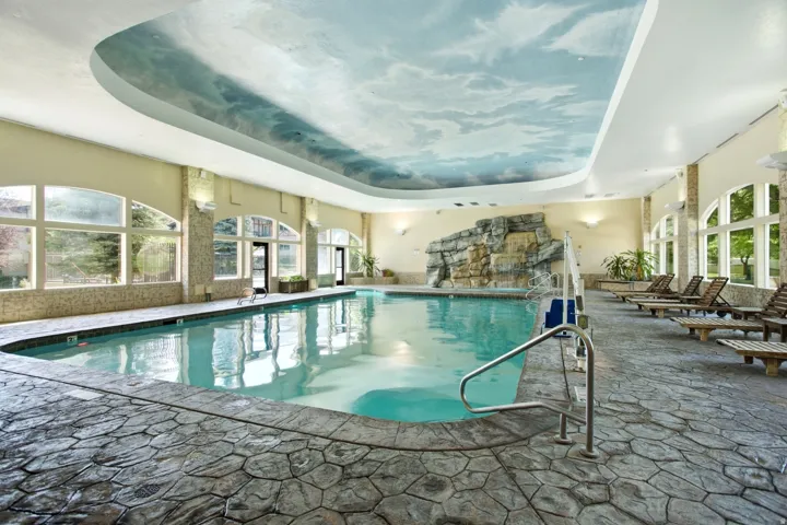 Indoor swimming pool with a stone-finish deck, rock waterfall feature, and a cloud-mural ceiling
