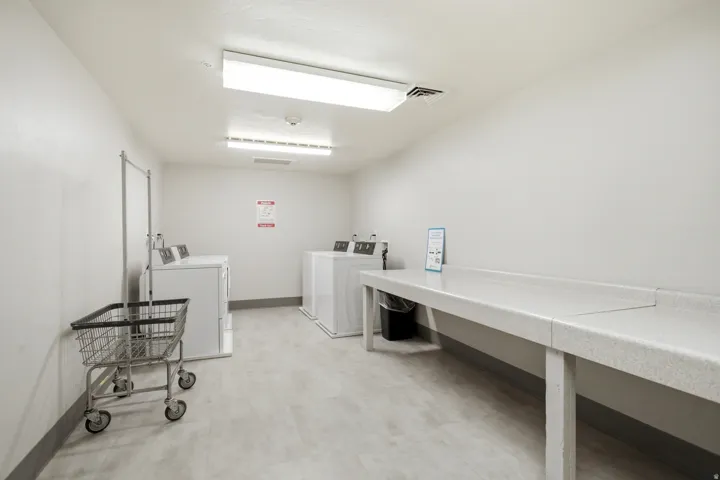Dedicated laundry room featuring commercial-grade washer and dryer units, generous countertop workspace with stainless steel sink and faucet, bright overhead LED lighting, and neutral tile flooring throughout