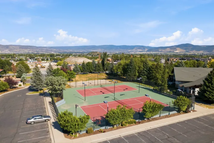 Two hard-surface tennis courts with red playing areas and green surrounds, enclosed by fencing, featuring multiple lighting fixtures