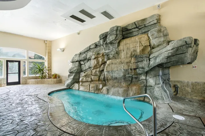Indoor aquatic recreation area featuring a resort-style lagoon-shaped pool with turquoise water, natural stone waterfall feature with textured rock formations, heated flooring throughout, recessed ceiling ventilation, arched architectural detailing, and d