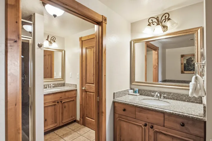 Dual-sink bathroom featuring warm wood cabinetry with ample storage, granite countertops, framed mirrors with integrated lighting, and tile flooring