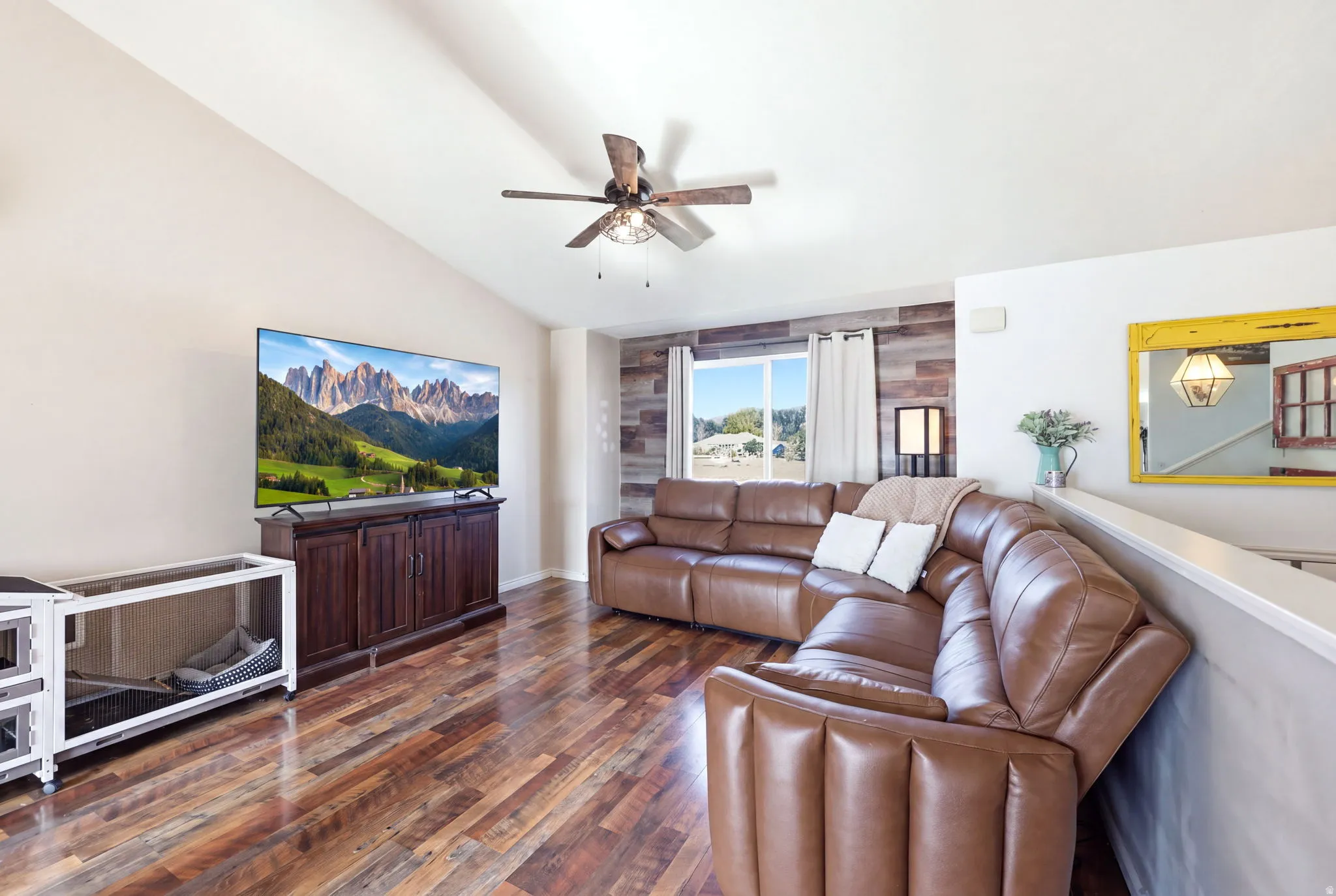 Spacious living room showcasing rich wood-finish flooring, a ceiling fan with integrated lighting, and large windows allowing natural light