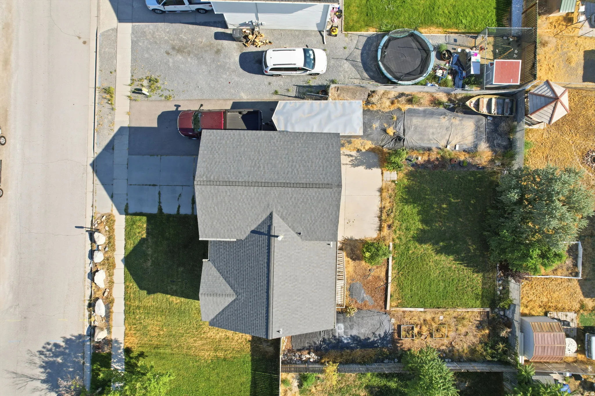 Aerial view showcasing a gray composite shingle roof with multiple peaks and angles, well-defined landscaping with mature trees and established lawn areas, concrete driveway, and adjacent single-family residential properties