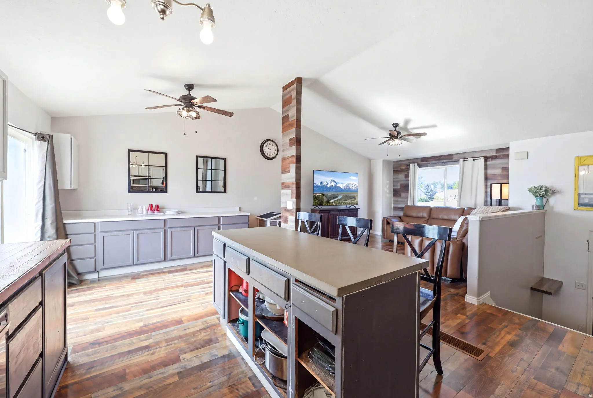Open-concept kitchen and dining area featuring a large center island with built-in shelving and wood-finish countertop, stainless steel appliances, custom cabinetry with brushed hardware, wood-finish flooring throughout, ceiling fans with lighting, and ab