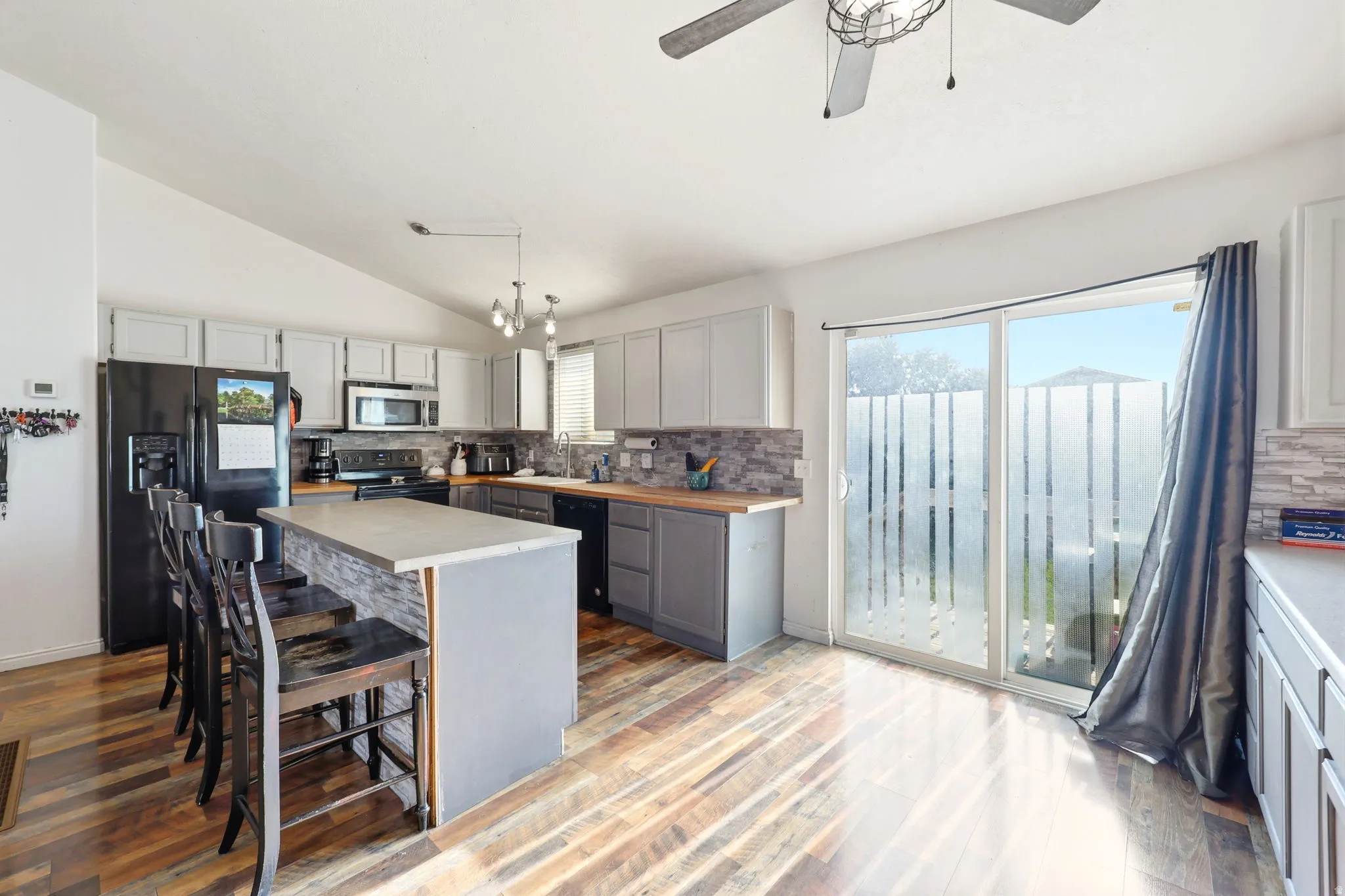 Modern kitchen and dining area featuring a large center island with white cabinetry and gray quartz countertops, stainless steel appliances including a black refrigerator, and pendant lighting