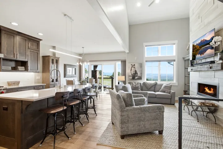 Vaulted ceiling great room featuring a stone fireplace, large windows, wood-finish flooring, a kitchen island with seating, and dark wood cabinetry