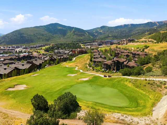 Sprawling golf course green with sand traps, surrounded by residential properties featuring varied architectural styles and mature trees, all set against a backdrop of distant mountains