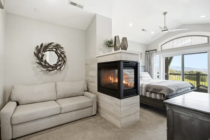 Luxurious primary suite featuring a double-sided linear fireplace with stone surround, plush seating area with neutral upholstery, soaking tub with dark finishes, and expansive glass doors opening to panoramic grounds