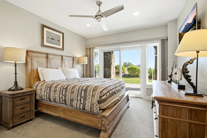 Spacious primary bedroom featuring a wood-frame bed, ceiling fan with recessed lighting, dual nightstands with task lighting, and direct access to a covered patio overlooking manicured landscaping