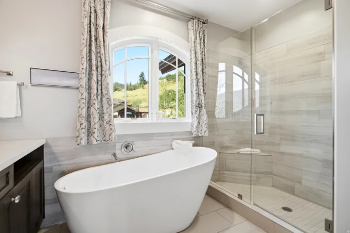 Luxurious primary bathroom featuring a freestanding oval soaking tub, frameless glass-enclosed walk-in shower with linear tile detailing, dual vanity with black cabinetry, and arched transom window framing natural light and outdoor scenery