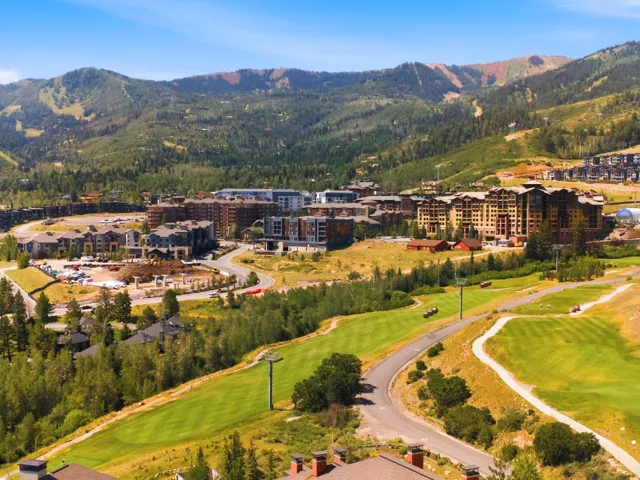 Expansive alpine resort community nestled in a mountain valley, featuring multi-story residential and hospitality buildings with warm-toned exteriors, manicured fairways and landscaped grounds, mature coniferous and deciduous trees throughout the property