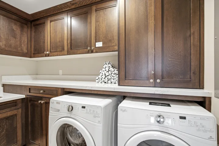 Dedicated laundry room featuring rich wood-finish cabinetry with ample storage, a quartz countertop workspace, and two full-size front-load appliances with modern control panels and stainless steel finishes