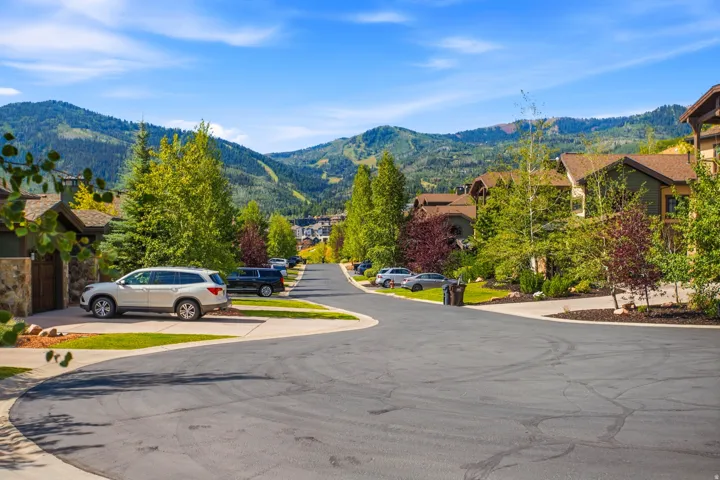 Peaceful residential street lined with mature trees and landscaped grounds, featuring well-appointed homes with stone and wood exteriors set against a backdrop of rolling, forested mountains