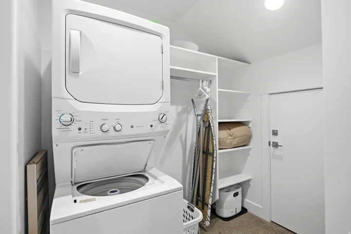 Dedicated laundry room featuring a stackable washer and dryer unit with electronic controls, open shelving for storage and organization, neutral flooring, and abundant natural lighting from recessed ceiling fixtures