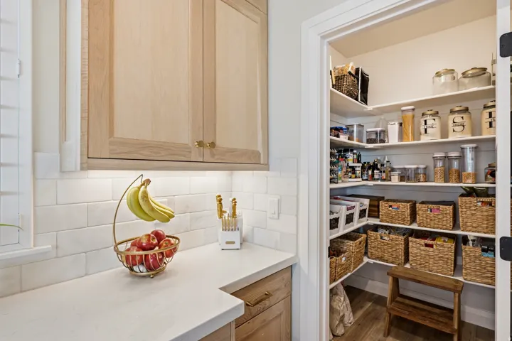 Spacious walk-in pantry with extensive shelving, complemented by light-toned upper cabinetry, white subway tile backsplash, and light-colored countertops