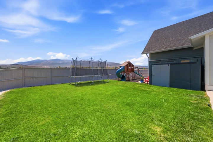 Expansive backyard oasis featuring a lush, manicured lawn with mature landscape borders, vinyl privacy fencing, a trampoline with safety enclosure, detached storage shed with dark exterior finish, and distant mountain vistas