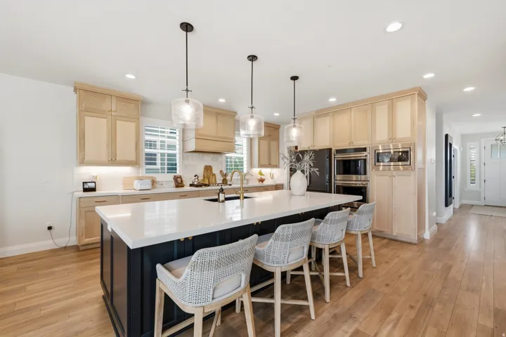 Expansive kitchen featuring a large central island with seating, light wood-finish cabinetry, built-in stainless steel appliances, pendant lighting, and wood-finish flooring