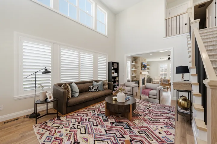 Two-story living area featuring tall windows with plantation shutters, wood-finish flooring, a carpeted staircase with black metal balusters, and an open entryway to an additional living space