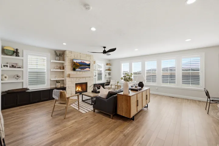 Spacious living room anchored by a stacked-stone fireplace with built-in shelving and cabinetry, wood-finish flooring throughout, abundant natural light from multiple shuttered windows, ceiling fan with recessed lighting, and a black upholstered sofa with