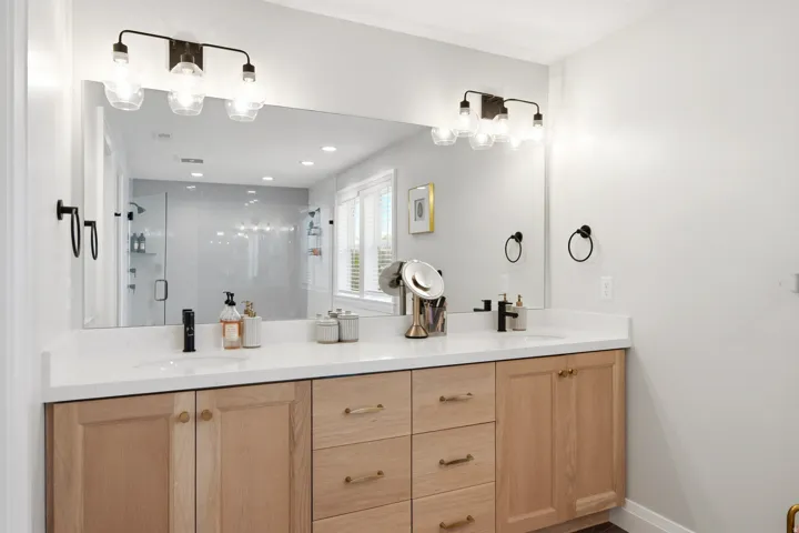 Spa-inspired primary bathroom featuring a dual-sink vanity with warm wood-finish cabinetry, white quartz countertop, and polished chrome hardware