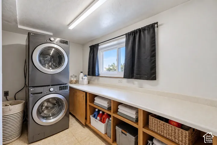 Laundry room featuring stacked washer and dryer unit, extensive solid-surface countertop, custom wood cabinetry, open shelving, and sliding window
