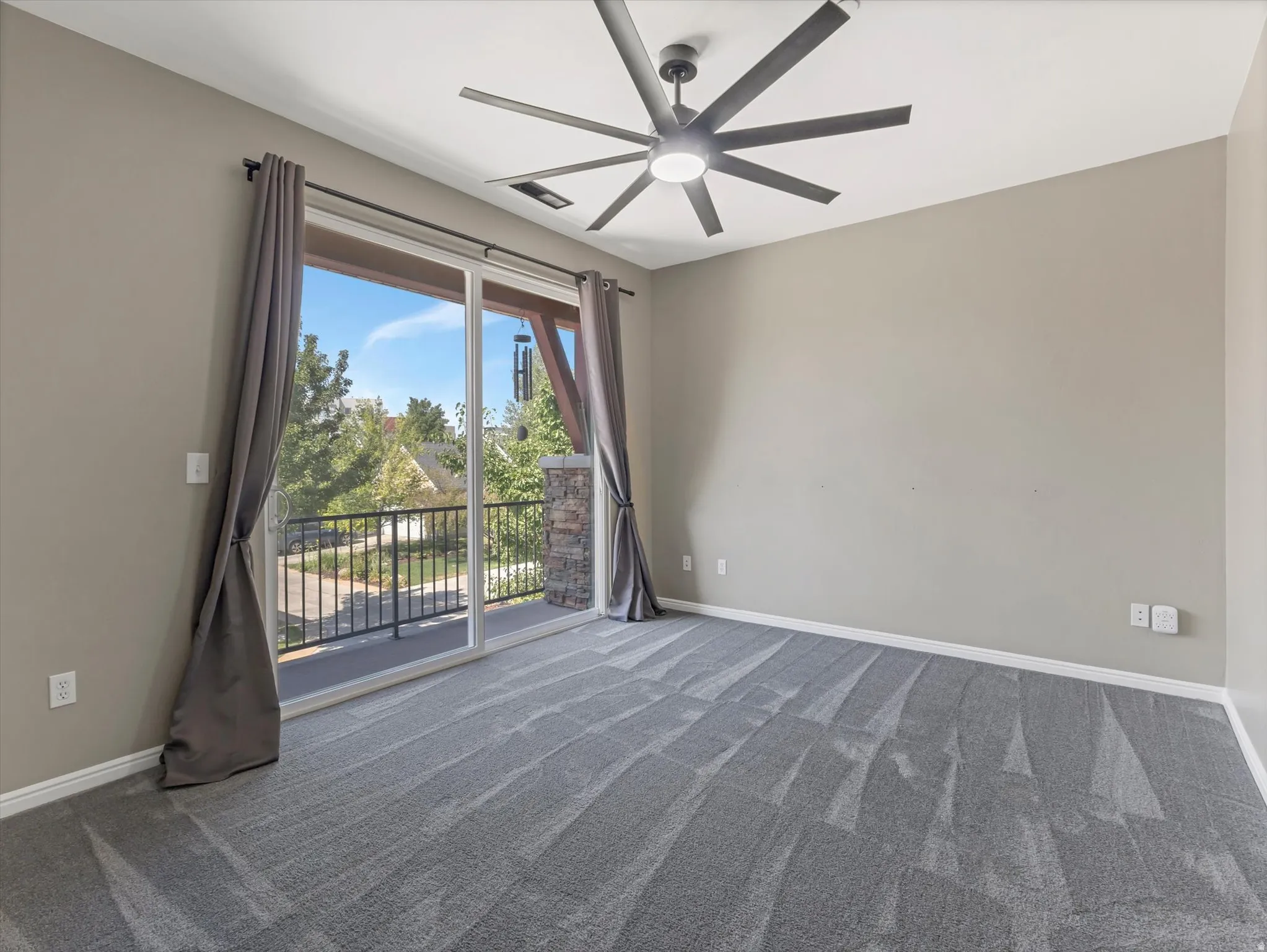Spacious room featuring neutral painted walls, gray patterned carpet, a modern ceiling fan, and large sliding glass doors opening to a private balcony with a stone column