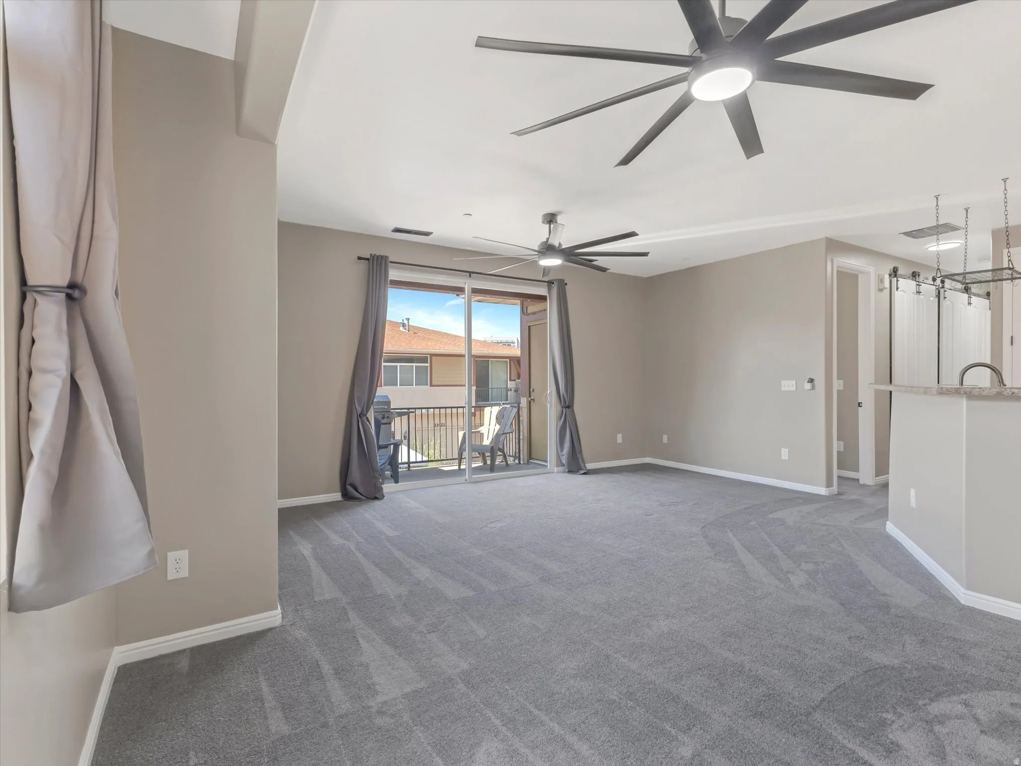 Spacious living area featuring neutral-toned walls, plush grey carpeting, two ceiling fans with integrated lighting, and a sliding glass door leading to a private balcony